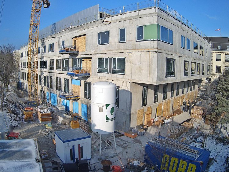 A multi-storey building in the shell with a concrete façade and windows already in place, surrounded by a building site featuring a crane, building materials and containers. In front of the building are construction vehicles and a large cylindrical silo, whilst trees and other buildings can be seen in the background.
