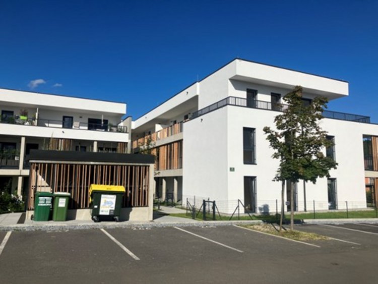Zwei weiße Gebäudeteile mit einem Mistplatz, Parkplätzen und einem Jungbaum davor