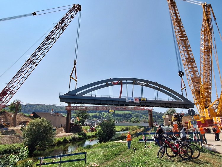 Zwei große Kräne heben eine Stahlbrücke über einen Fluss.