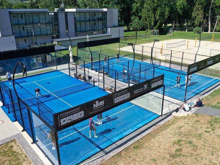 Two padel courts in use, with a beach volleyball court next to them and a three-storey clubhouse behind them.