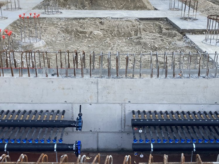 Bauplatz mit einem an einer noch nicht fertiggestellten Betonwand befestigten Verteilerbalken als Wandverteiler im Verteilerraum mit bereits überdeckten Anbindeleitungen sowie Pfahlköpfen. 