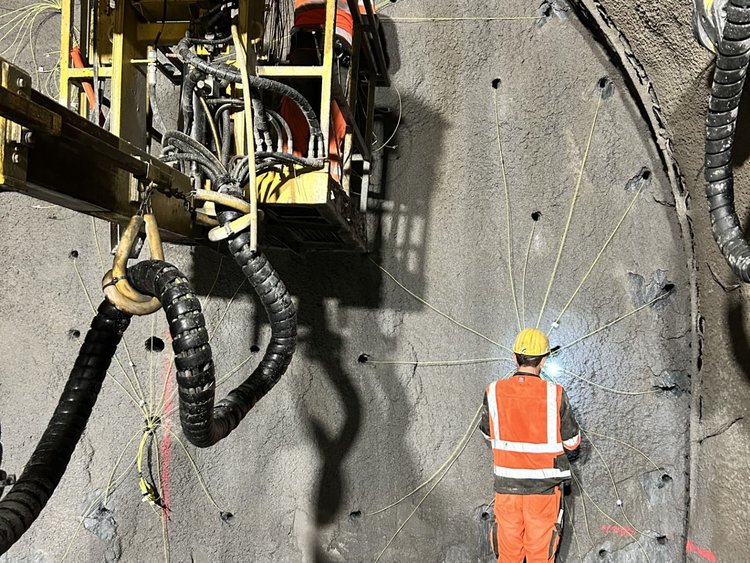 Staff in protective clothing are attaching detonating cords to the tunnel wall. One is standing on the ground, a second on a lifting platform