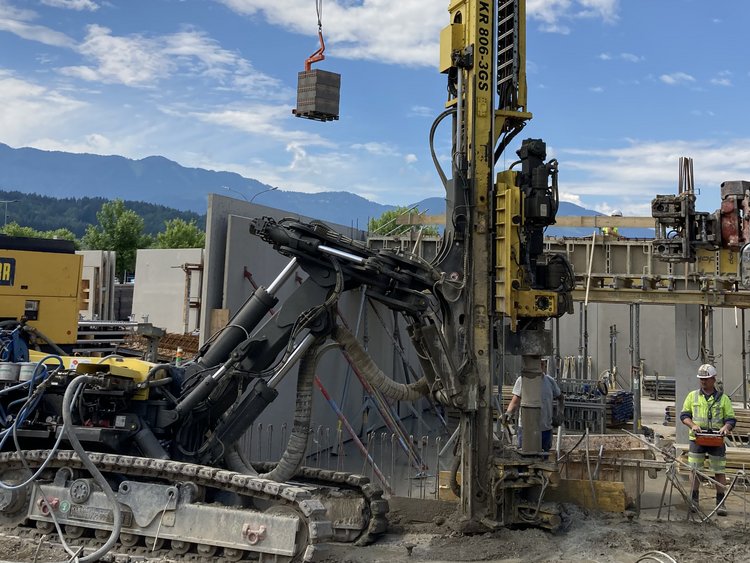 Baustelle mit einem Raupenbohrgerät im Einsatz. Zwei Männer bedienen das Gerät. Im Hintergrund Bäume und ein Gebirgszug.