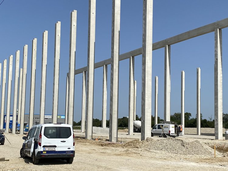Bauplatz mit ersten Pfeilern für den Bau einer Halle, rechts im Bild verschiedene Baumaterialien, daneben steht ein Auto, rechts im Bild weitere Baufahrzeuge