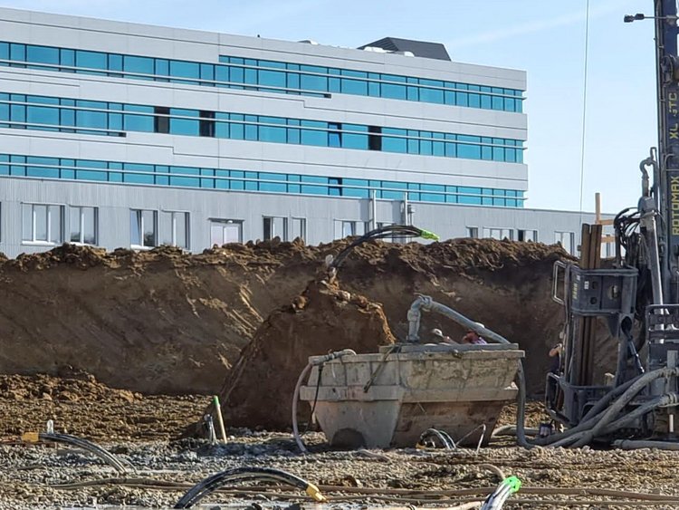 Im Vordergrund ein Bauplatz mit einem Geothermie-Bohrgerät und einer Absetzmulde, aus dem Boden ragen Leitungen. Im Hintergrund das bereits bestehende Gebäude der FH Campus Wien.