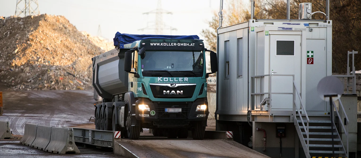 Foto: Lastwagen der Koller GmbH bei der Einfahrt ins Recycling Center Himberg auf der Waage; daneben Bürocontainer mit teilweise geschlossenen Jalousien, Türe und Scheinwerfern am Dach, im Hintergrund Schuttberge und Strommasten