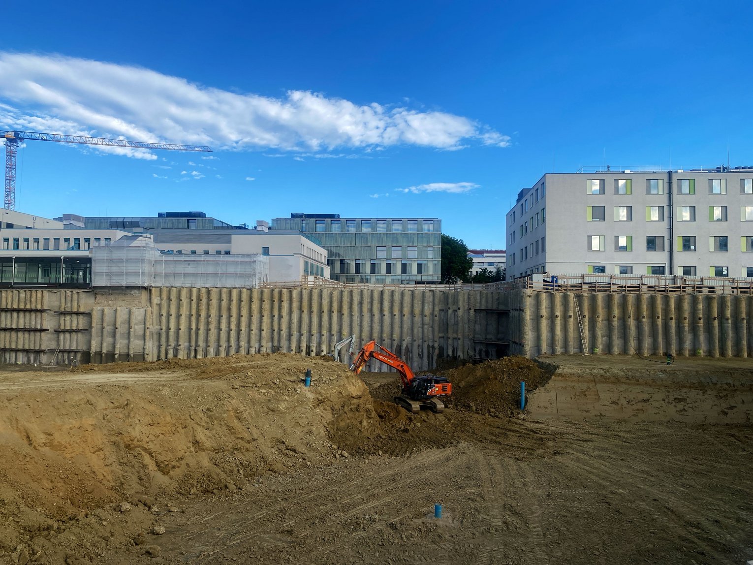 Construction site with an orange excavator digging in a sandy area, surrounded by a tall concrete sound barrier. Modern buildings and cranes are visible in the background under a clear blue sky.